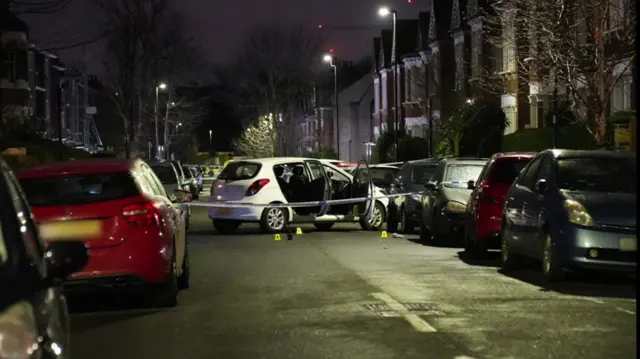 A white car with its doors open in a street filled with parked cars, wth police tape across it and cones on the road