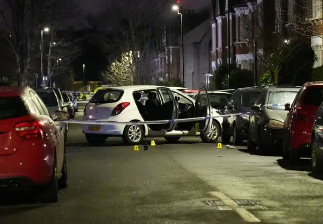 A white car, with its doors open, sits in the middle of a street with a police cordon around it