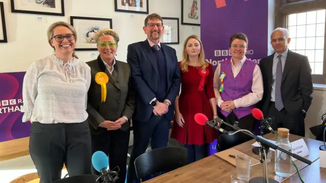 Four women and two men, some wearing political rosettes, standing behind a table on which microphones are visible