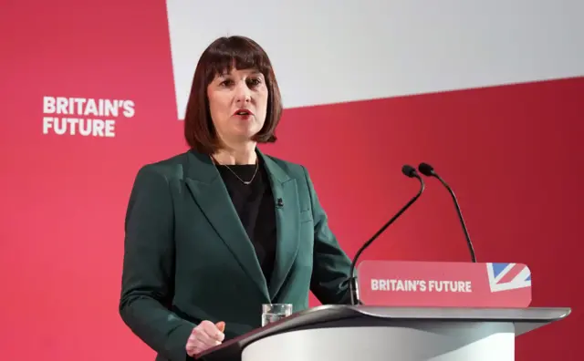 Shadow Chancellor Rachel Reeves speaking into a microphone at Labour Party Business Conference, with a red background