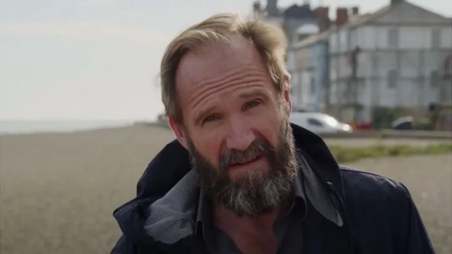 A close of up Ralph Fiennes with some buildings and a beach in the background, from his movie ‘Coast’