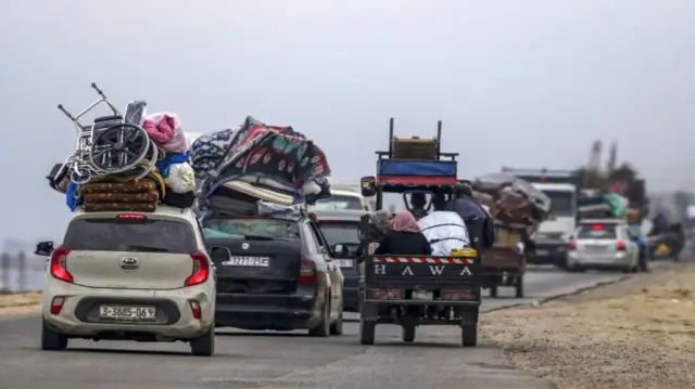 Several cars are seen, some packed with belongings on their roofs, including one with a wheelchair. A tuk tuk is carrying two women in hijab.