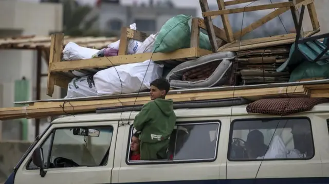 A boy leans out of a people carrier window, looking into the distance. Another boy is also looking out the same window at something. The roof of the van is piled with belongings.