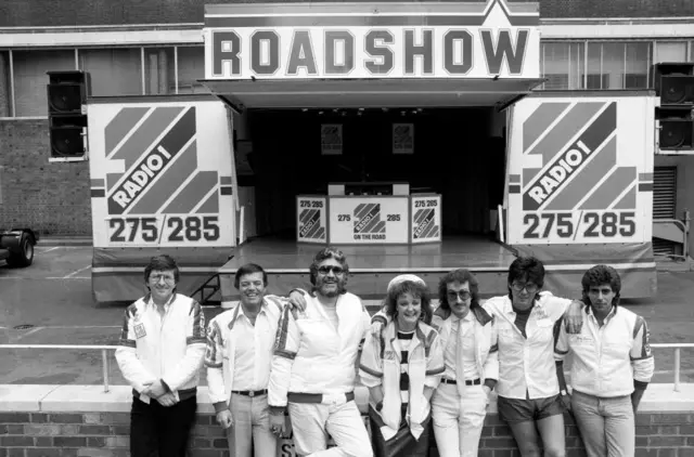 Radio One DJ's Simon Bates, Tony Blackburn, Dave Lee Travis, Janice Long, Steve Wright, Mike Read and Gary Davies standing in front of the Roadshow Studio