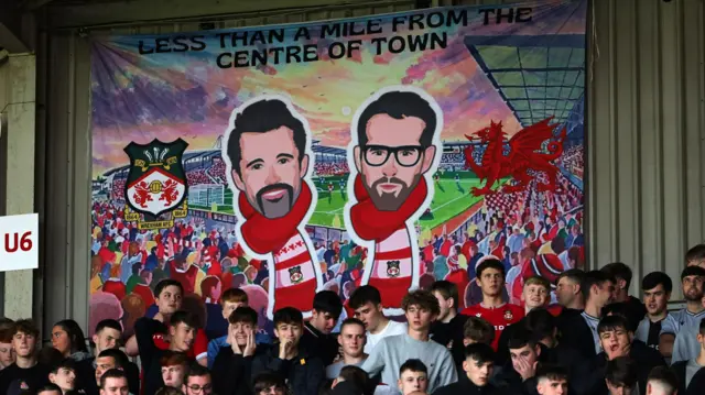 Wrexham fans in front of a banner of their club's owners, Hollywood stars Rob Mcelhenney and Ryan Reynolds