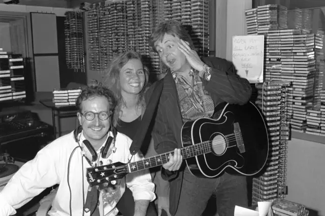 Paul McCartney, DJ Steve Wright and his production assistant Dianne Oxberry in a radio studio standing in front of cassettes