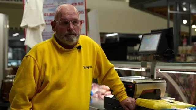 A man stood behind a weighing scale and till in a yellow jumper