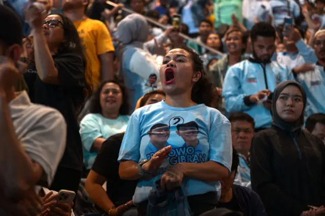 A woman wearing a t-shirt with Prabowo and his running mate cheers during his address, with a crowd of people around her