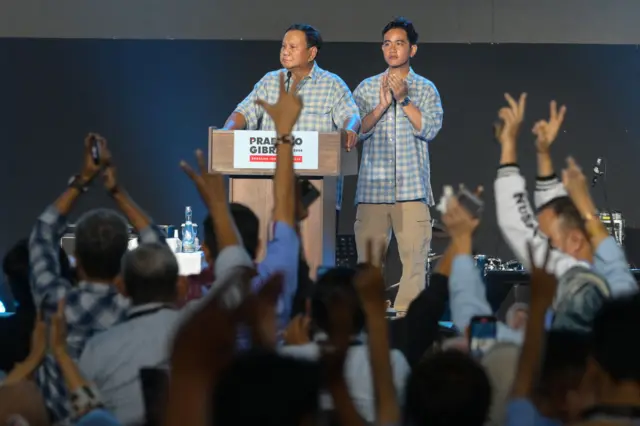 Prabowo Subianto speaking into a mic at a dais, while his running mate Gibran Rakabuming Raka stands behind him in front of a crowd of supporters