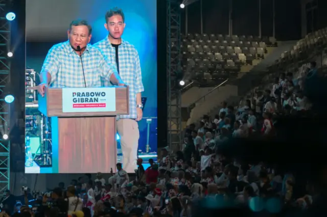 A large screen projects an image of Prabowo and his running mate, in front of a crowd of his supporters seated in what looks like a stadium