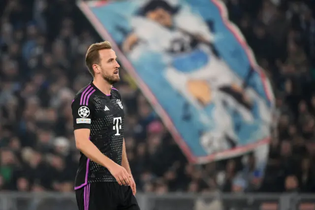 Kane stands and stares into space as Lazio fans fly a flag behind him.