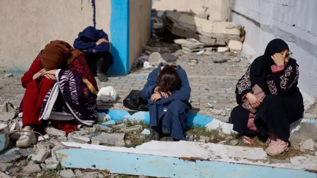 People rest next to damaged buildings, as Palestinian arrive in Rafah after they were evacuated from Nasser hospital in Khan Younis due to the Israeli ground operation