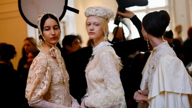 Three models get ready backstage to present creations by Turkish designer Bora Aksu. They are dressed in white, lacy dresses and Victorian-era hats