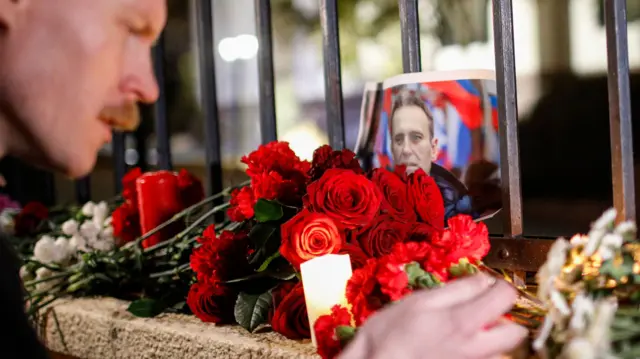 A man lights a candle on a wall next to flowers and a picture of Russian opposition leader Navalny