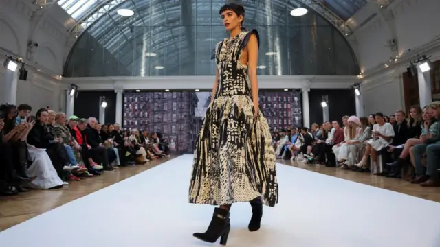 A model wears a black and white dress with black boots while walking the runway during a show at London Fashion Week