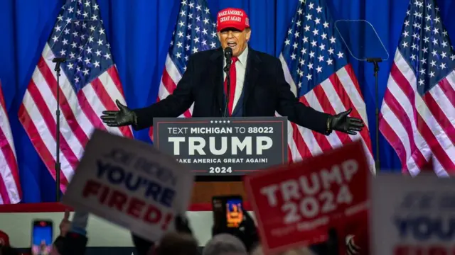 Former president Donald Trump speaks at a lectern with a red MAGA hat