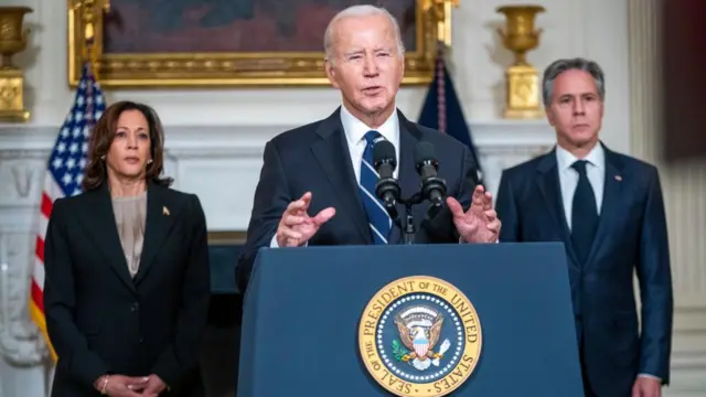 Joe Biden speaks at a lectern at the White House. Behind him stand Harris and Blinken