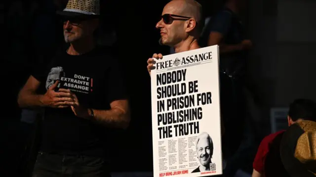 A man in sunglasses holds a poster designed to look like a newspaper article that reads: Nobody should be in prison for publishing the truth