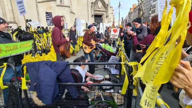 People playing musical instruments outside court in central London with flyers and protest signs in support of Julian Assange