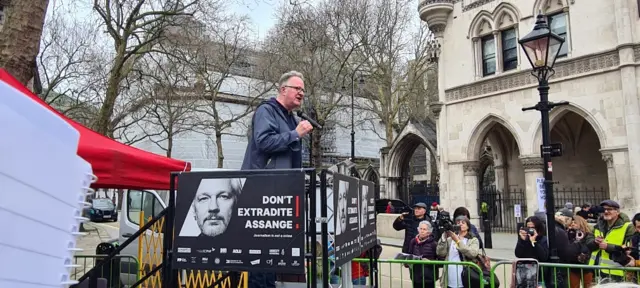 Tim Dawson from the International Federation of Journalists stands on a temporary stage holding a mic and giving a speech to the crowd.