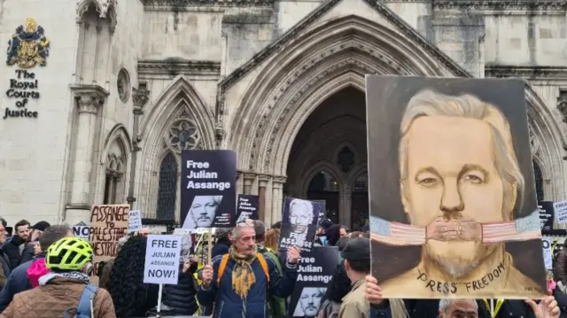 Protestors stand outside court room holding posters and signs that read "Free Julian Assange"