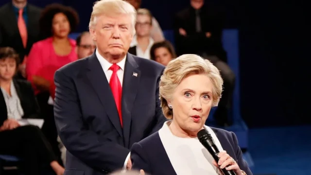 Clinton speaks into a microphone as Trump stands behind, watching her.