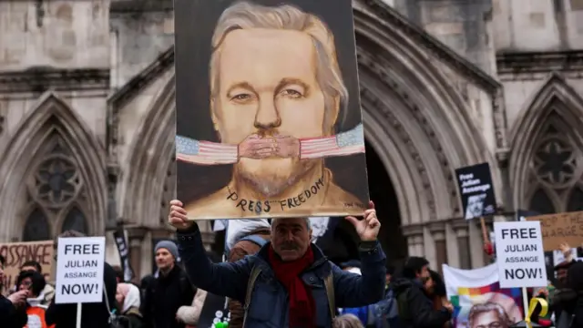 A crowd outside the Royal Court of Justice hold various banners in support of Assange. In the centre, a man holds overhead a painting of Assange's mouth being covered by hands emerging from US flags