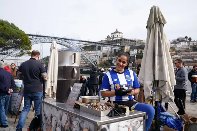 A porto fan stands in her club top behind her roasted chestnut stand at the riverside in Porto.