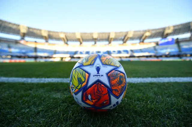 The UCL match ball stands on the turf at Stadio Armando Maradona.