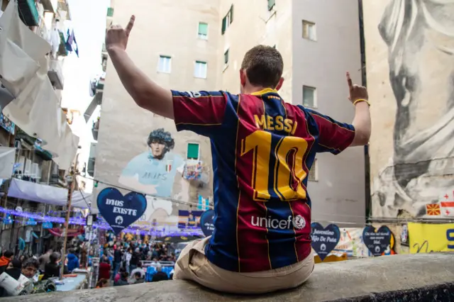 A fan with a 'Messi' Barcelona shirt sits on a wall in front of the Maradona mural in Naples.