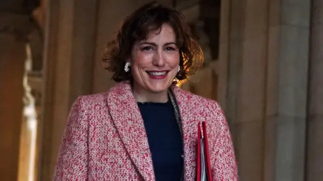 Victoria Atkins, wearing a red tweed jacket, walks through a stone archway, carrying a red folder