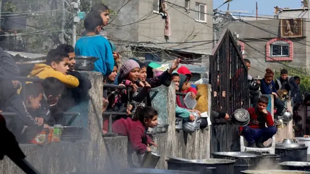 Palestinian children hold bowls, pans and tins through a metal gate