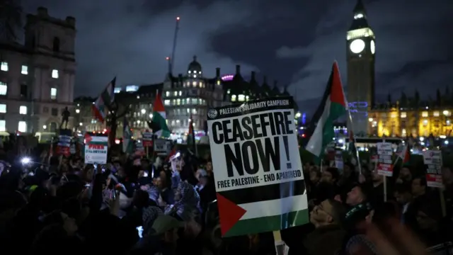 A "ceasefire now" sign is in focus, in front of a crowd outside Westminster