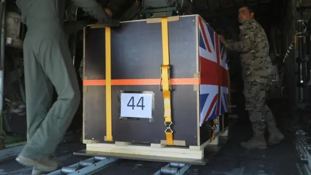 Two people in military uniform load a large box branded with a Union Jack onto a military plane