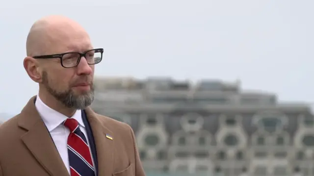 Arseniy Yatsenyuk wearing a brown blazer, white shirt, and red and white striped tie, as he speaks, with Kyiv's skyline in the background