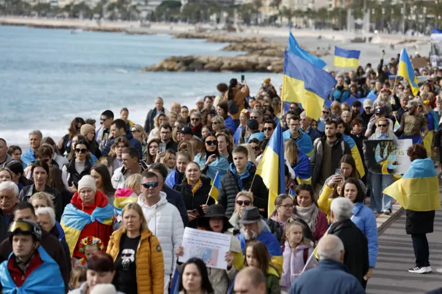 A crowds of people march along the coastline in Nice holding Ukrainian flags