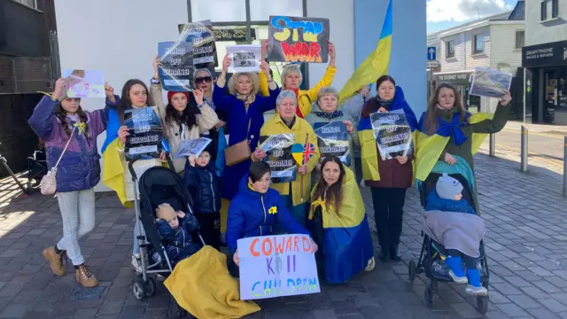 A group of Ukrainian refugees, holding signs and draped in Ukraine flags, pose for a photo