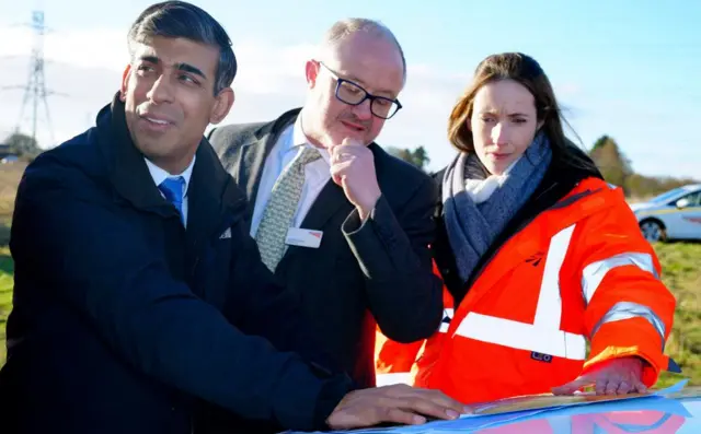 Sunak,  Andrew Haines and Anna Weeks inspect paper plans, which are spread out on the bonnet of a car in a grassy field