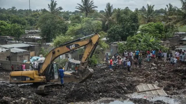 Residents stand next to an excavator working at the site of a dump collapse in a district of Maputo on February 19, 2018.