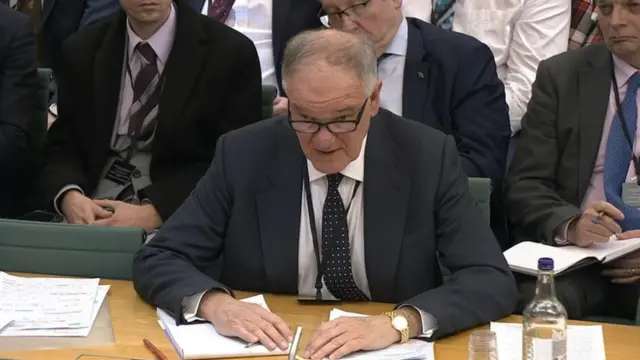 Staunton, wearing a suit, sits at a table, speaking to the committee