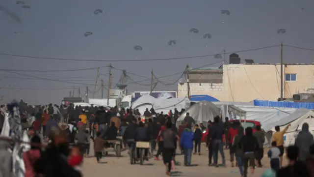 Hundreds of people mill about a road lined with tents. In the air above, ten parachutes float towards the ground