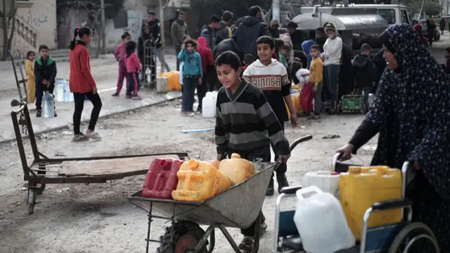 A boy transports jerry cans of water in a wheelbarrow. Behind him, a crowd surrounds what looks like a petrol truck presumably stocking water