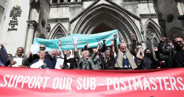 Sub-postmasters hold their hands in the air in front of a red banner with the words “Support our sub-postmasters”