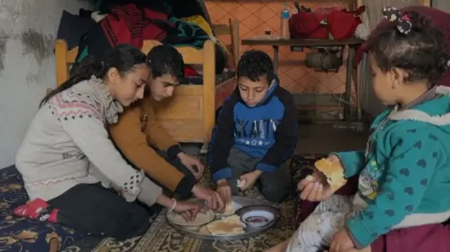 Malak and three other children sit on the floor together sharing food from a tray