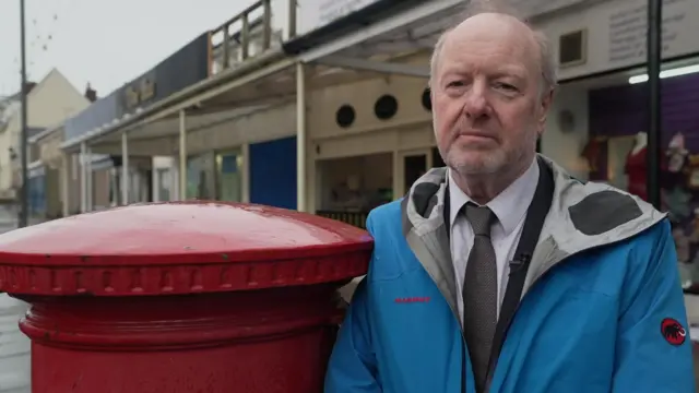 Alan Bates wearing a light blue jackets leans against a red Post box