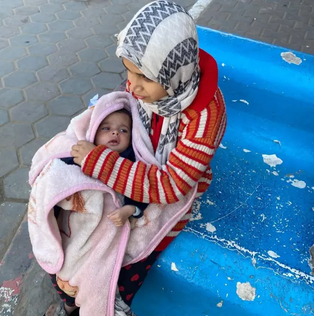 A young girl holds her baby brother tightly outside Al-Aqsa Martyrs Hospital in Deir al Balah