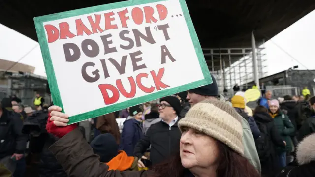 Woman protesting outside Senedd with a sign that reads: "Drakeford doesn't give a duck"