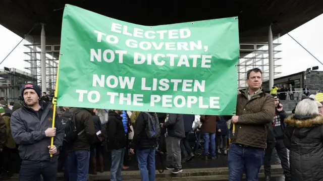 Two male protestors hold up sign that reads "Elected to govern not dictate. Now listen to the people"