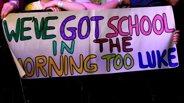 Children hold sign reading "We've got school in the morning too, Luke"