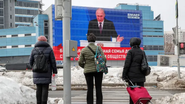 People stand in front of a large screen that shows a live broadcast of the Russian President Vladimir Putin delivering his annual address